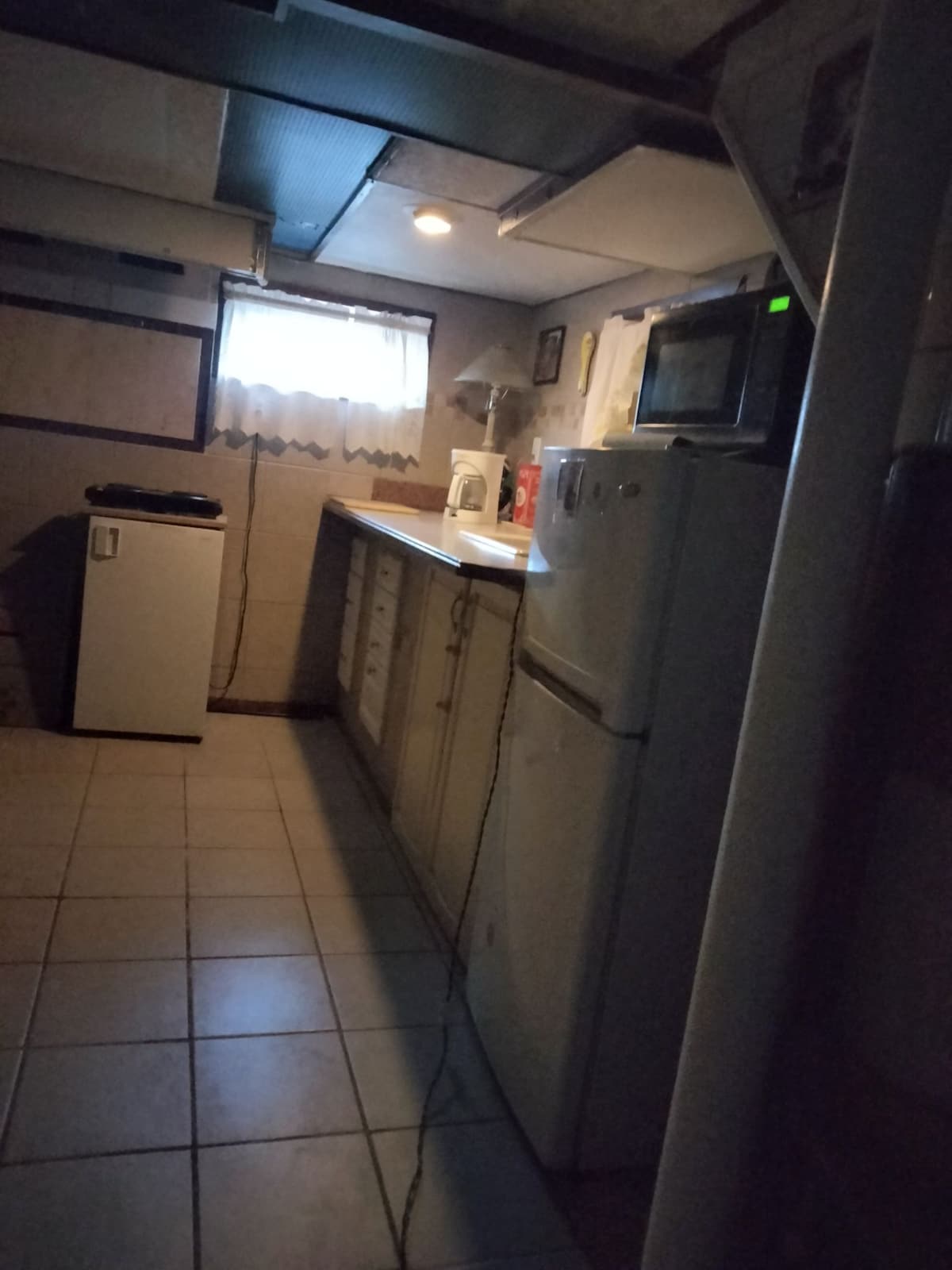 The kitchen area is illuminated by overhead lighting, showcasing a refrigerator and a compact freezer. A countertop with cabinets runs along one wall, and a microwave is positioned for easy access. A window with curtains allows natural light to enter the space.