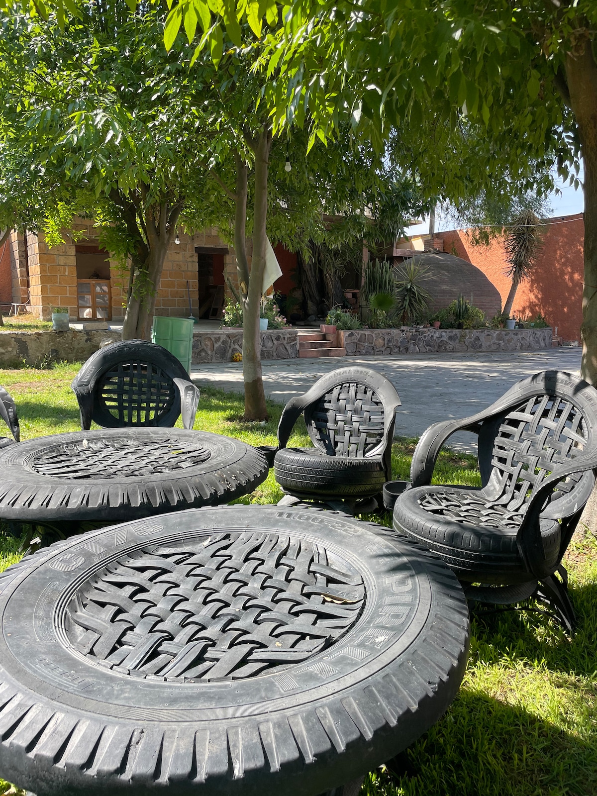 An outdoor seating area features several comfortable chairs and a round table made from recycled tires, surrounded by vibrant green grass and trees. A part of the property is visible in the background, showcasing a serene and inviting atmosphere.