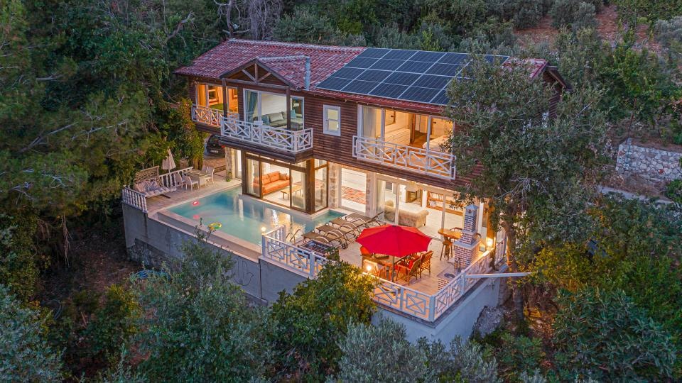 A two-story villa is surrounded by greenery, featuring solar panels on the roof. The outdoor area includes a swimming pool, lounge chairs, and a red umbrella over a dining table. Soft lighting enhances the inviting ambiance during dusk.