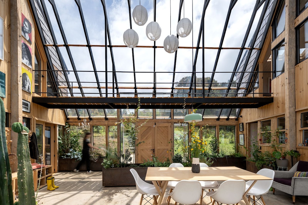 A spacious greenhouse features large glass panels allowing natural light to fill the room. A wooden dining table is surrounded by modern chairs. Lush greenery is placed throughout the space, with decorative lanterns suspended from the ceiling, creating a serene indoor setting.