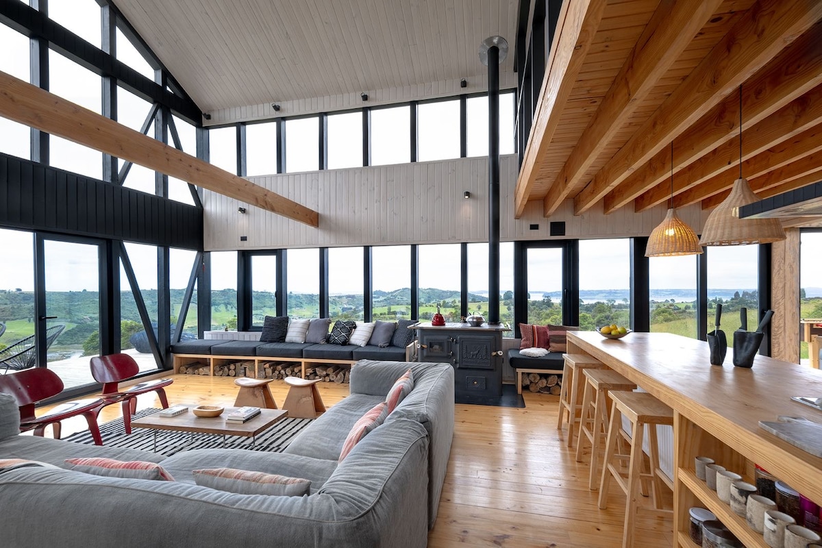 A spacious living area showcases high ceilings with exposed wooden beams. Large windows provide panoramic views of the surrounding landscape. A comfortable gray sectional sofa is positioned opposite a wooden dining table and bar area, creating an inviting space for relaxation and gatherings.