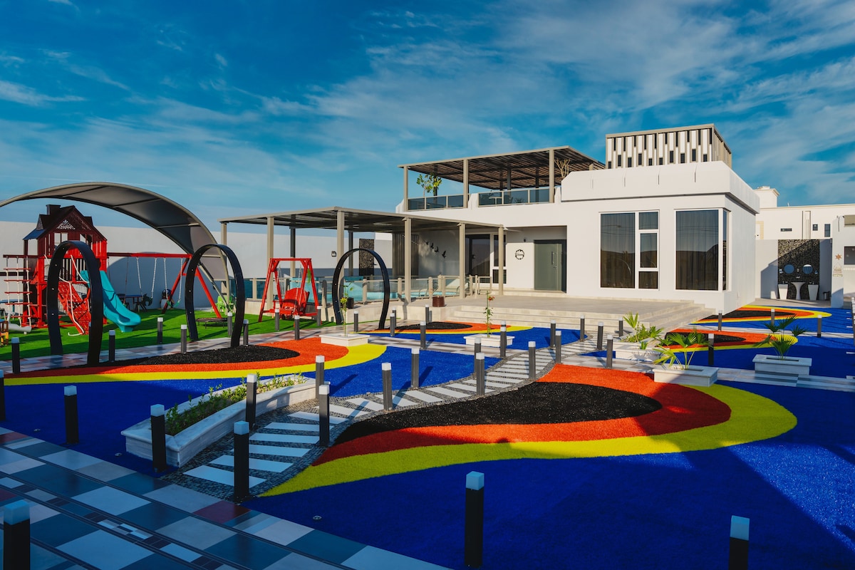 An outdoor recreational area features colorful surfaces in blue, red, and yellow, with a playground equipped with swings and slides. A spacious building with large windows is visible in the background, providing a vibrant and engaging environment for families.