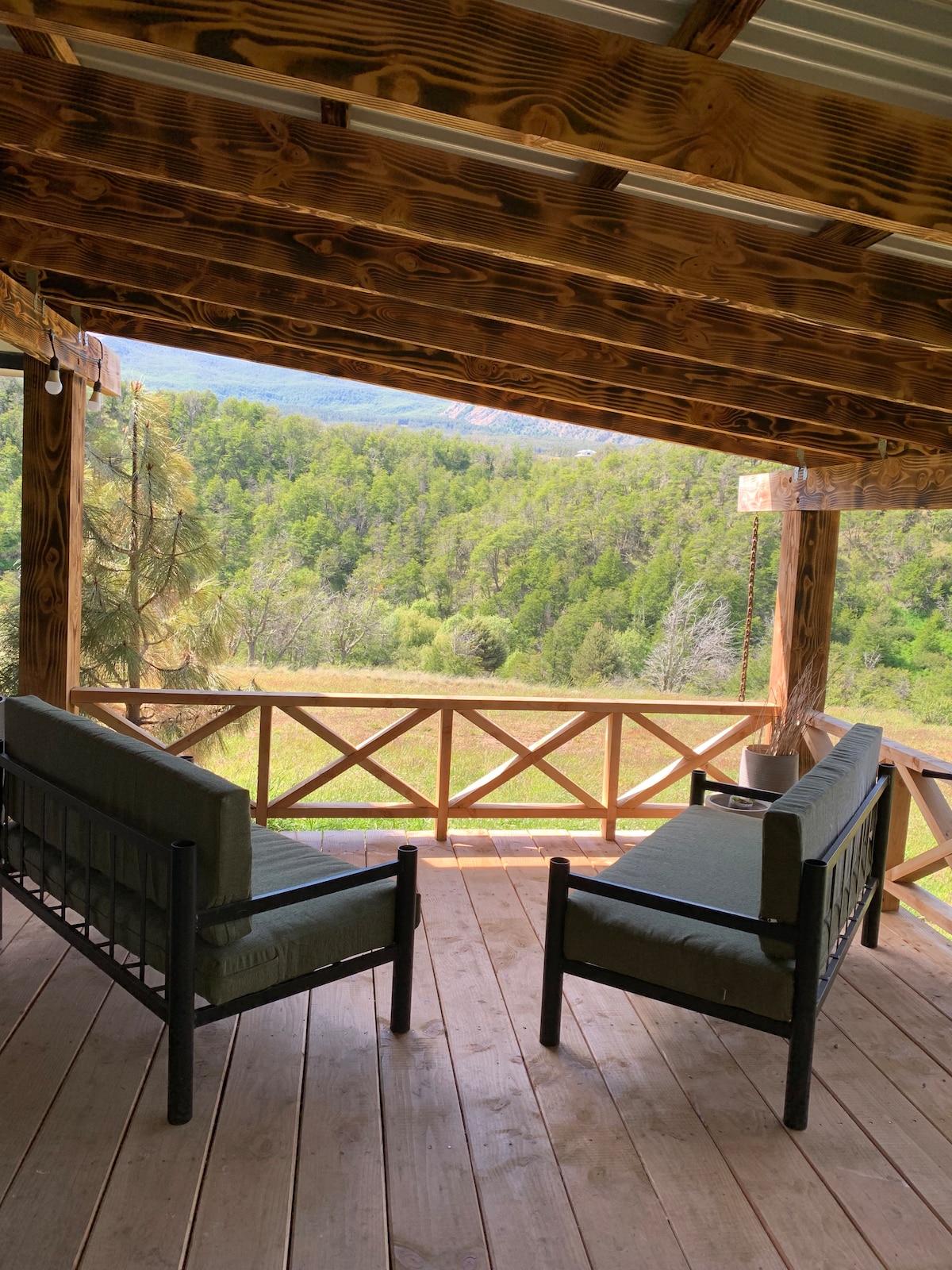 A covered terrace features two comfortable seating areas facing a scenic view of lush greenery and rolling hills. The wooden structure and flooring create a rustic ambiance, while the open space invites relaxation and enjoyment of the surrounding landscape.
