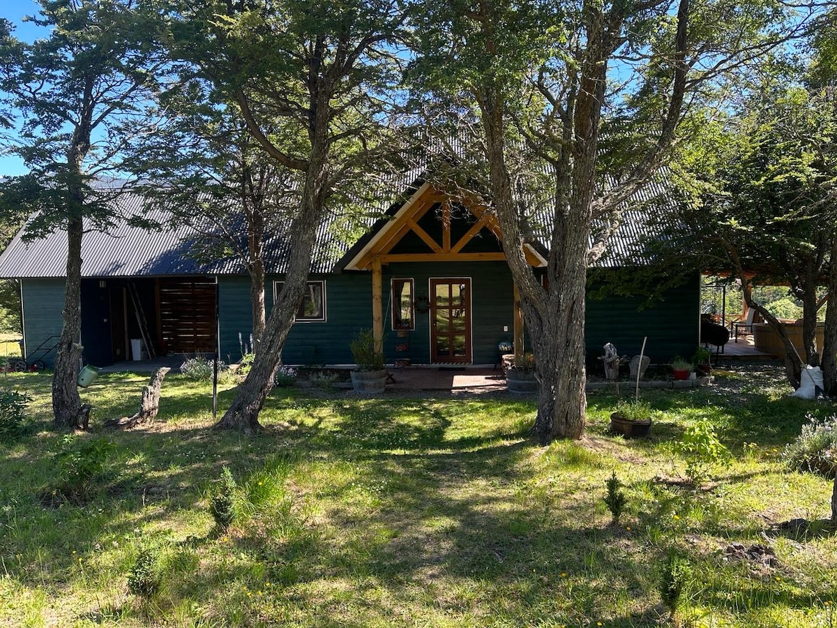 A welcoming house is set within a natural landscape, framed by trees. The entrance features a wooden door and large windows, inviting light into the interior. A manicured path leads from the foreground to the home's front, creating a serene atmosphere in the surroundings.