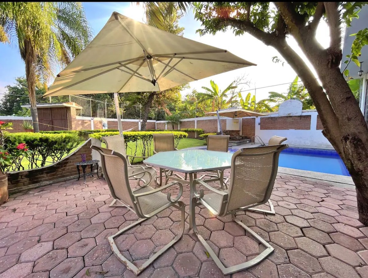 A patio area is presented with a round glass table surrounded by four metal-framed chairs. A beige umbrella provides shade. Lush greenery and a serene pool area are visible in the background, contributing to an inviting outdoor setting.