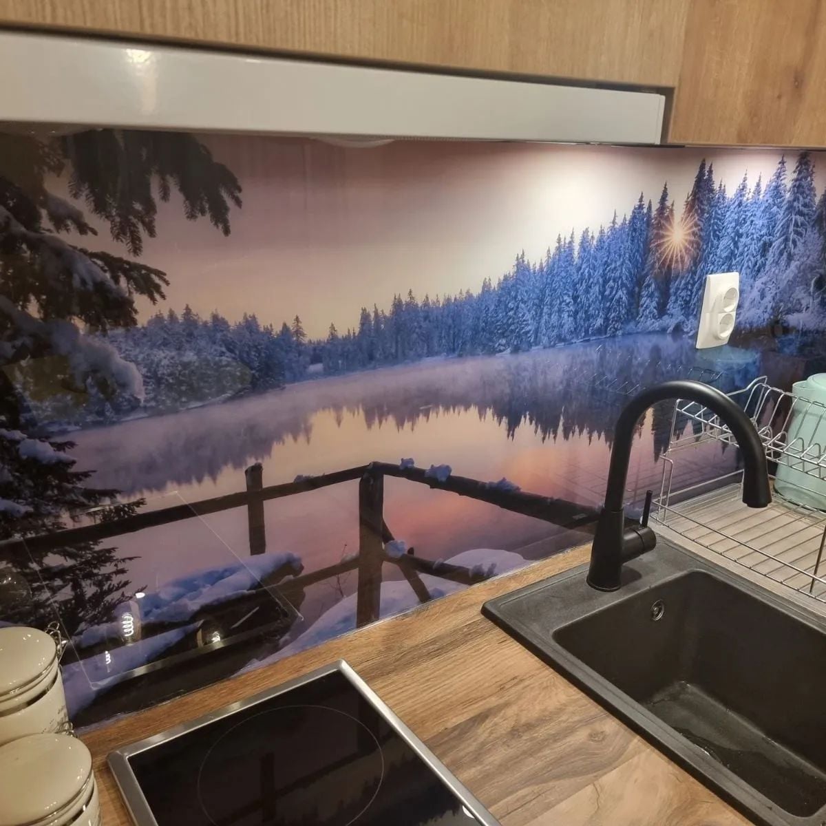 A serene winter landscape is depicted on the kitchen backsplash, featuring a tranquil lake surrounded by snow-covered trees. The scene captures the soft hues of a sunset, with gentle reflections in the water. A modern black sink and stovetop are visible in the foreground.