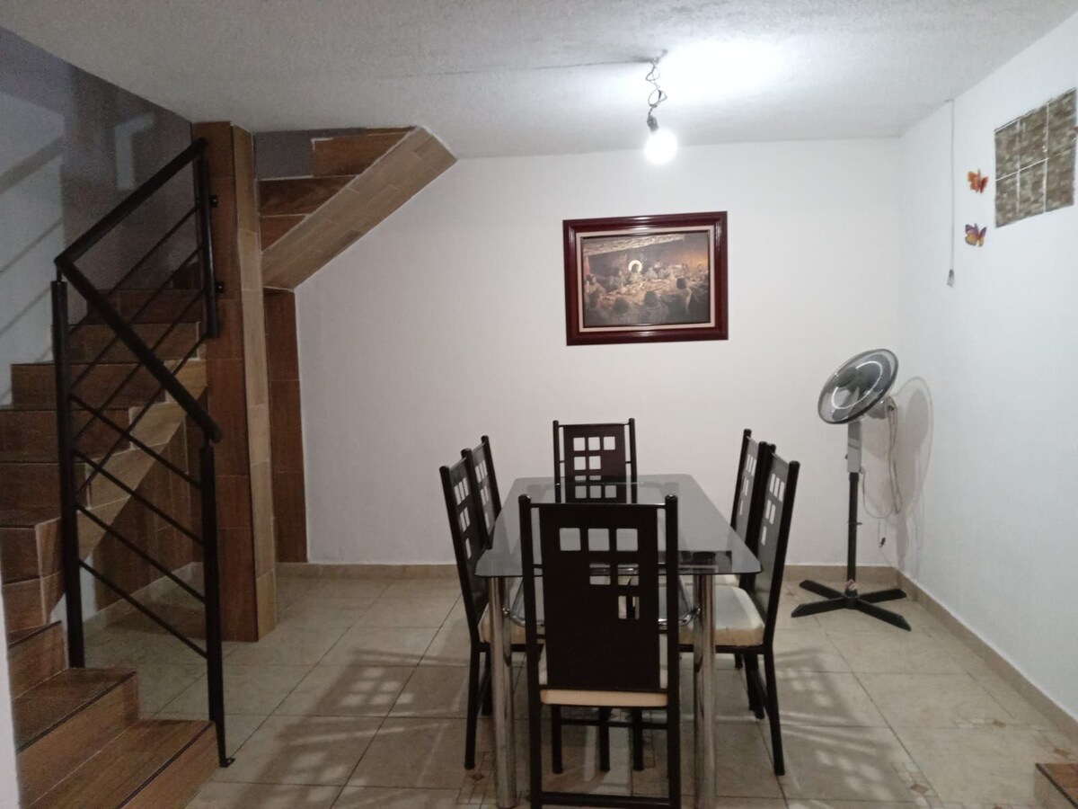 An elegant dining area is presented with a glass-topped table surrounded by six chairs. A light fixture hangs from the ceiling, illuminating the space. A staircase with wooden steps is visible in the background, complemented by a framed artwork on the wall.