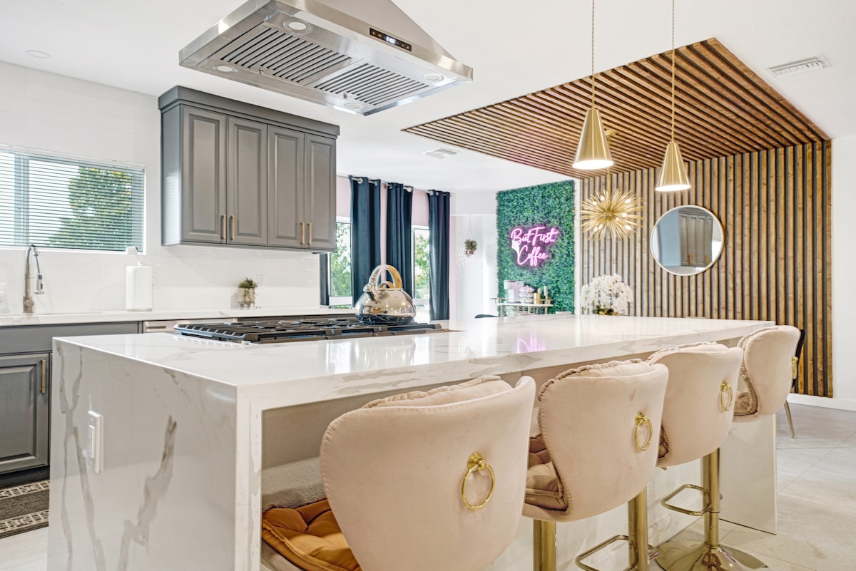 A modern kitchen features an expansive quartz island with four upholstered seats. Stylish cabinetry in gray contrasts with a wood-accented ceiling and decorative plants. A neon sign and a circular mirror add artistic touches, while natural light filters in through a nearby window.