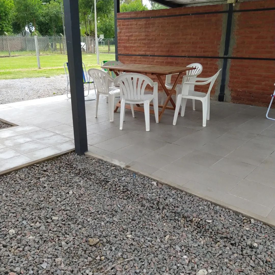 A covered outdoor area features a wooden table surrounded by four white plastic chairs. The flooring is made of gray tiles, complemented by a gravel section. Green trees are visible in the background, enhancing the open space.