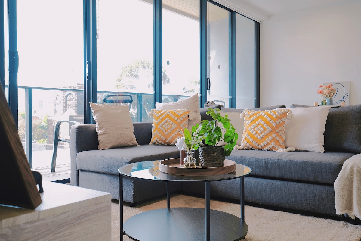 A comfortable living area is presented, featuring a gray sofa adorned with patterned yellow and white cushions. A round glass coffee table sits at the center, accompanied by a potted green plant. Large sliding glass doors provide access to a balcony, allowing natural light to brighten the space.