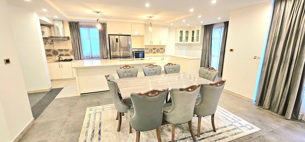 A dining area is featured with a large, rectangular table surrounded by eight upholstered chairs. A light-colored area rug lies beneath the table, and large windows allow natural light to fill the room. Stylish cabinetry and modern appliances are visible in the open kitchen space.