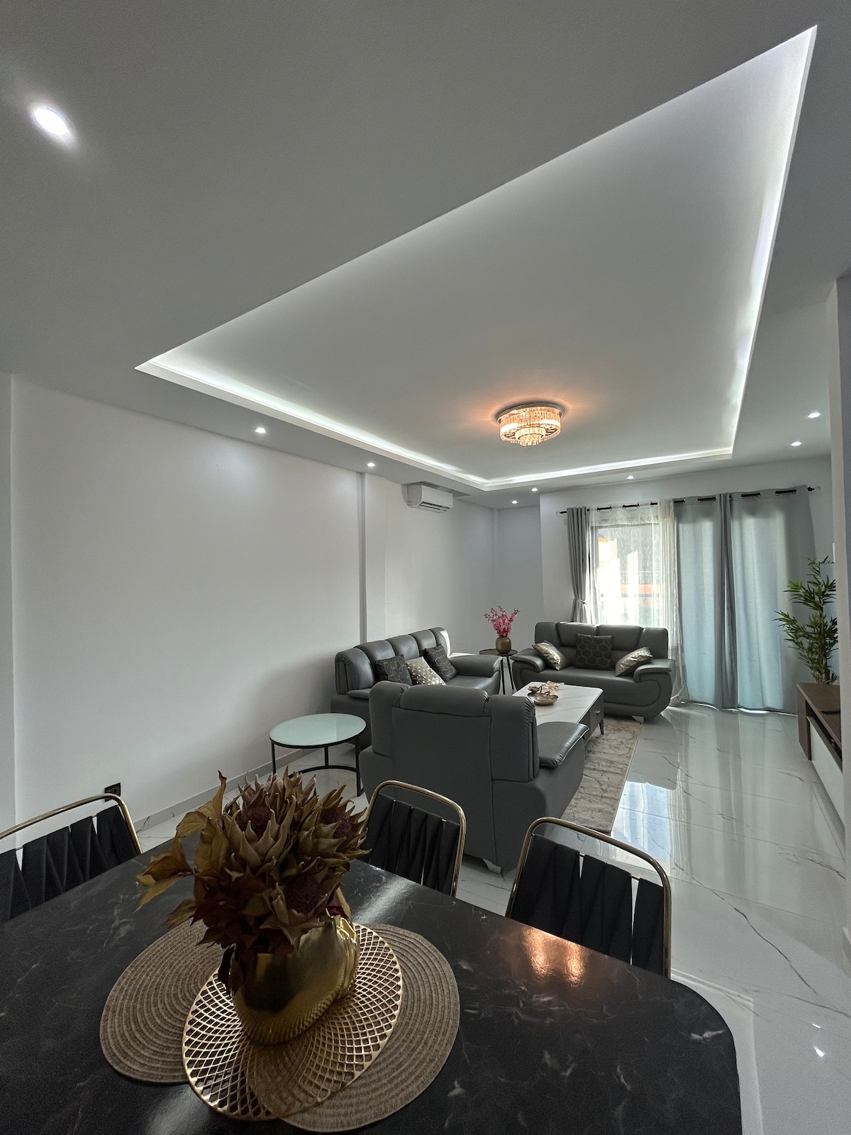 A contemporary living space is showcased, featuring a sectional sofa and sleek armchairs arranged around a coffee table. Natural light enters through large windows, complemented by sheer curtains. A dining area with a decorative centerpiece is visible, creating a sense of comfort and openness.