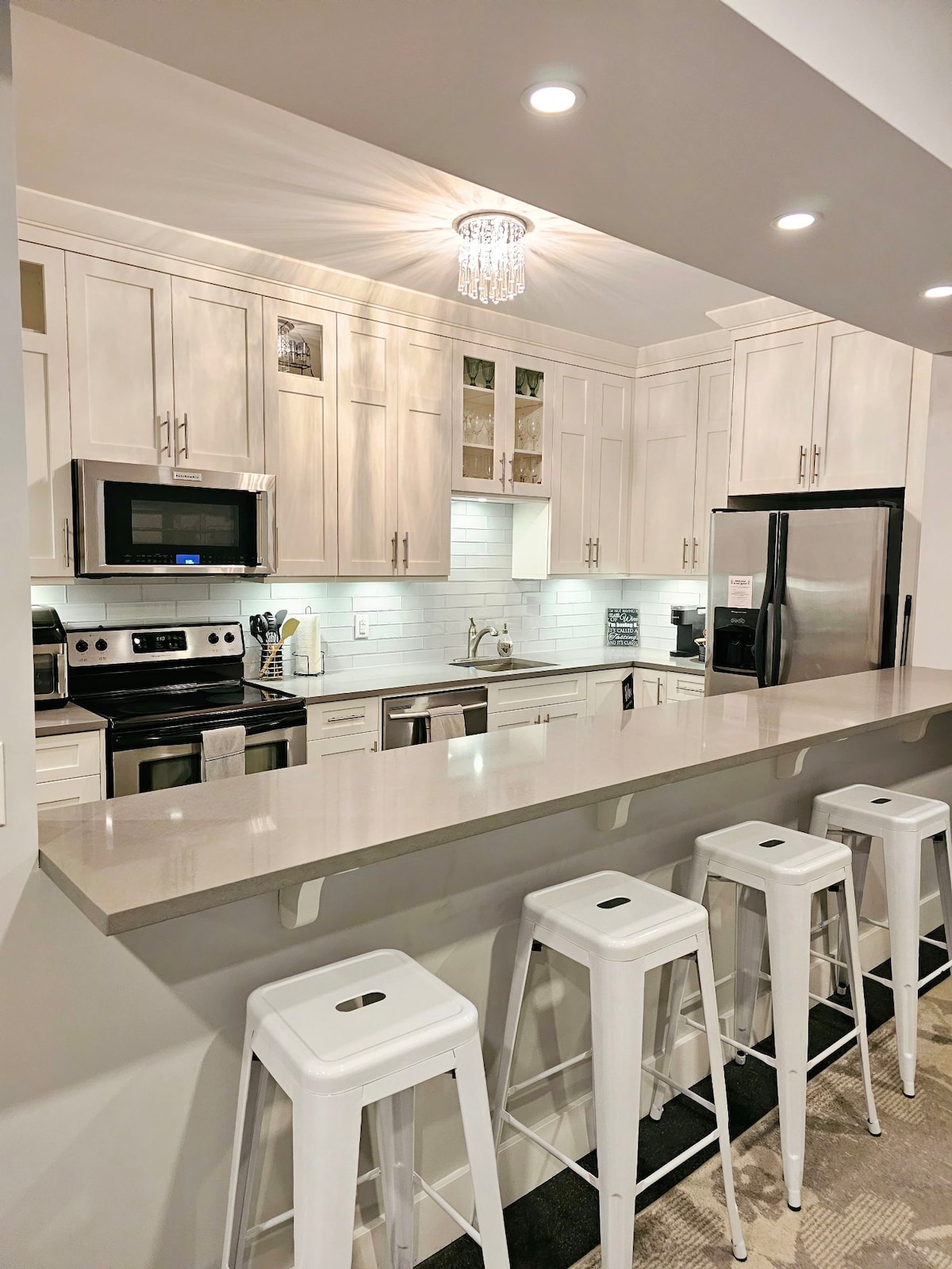 The modern kitchen features light cabinetry and a spacious countertop with four bar stools. Stainless steel appliances, including a microwave and an oven, are included. Subtle under-cabinet lighting highlights the stylish backsplash, enhancing the functional design of the space.