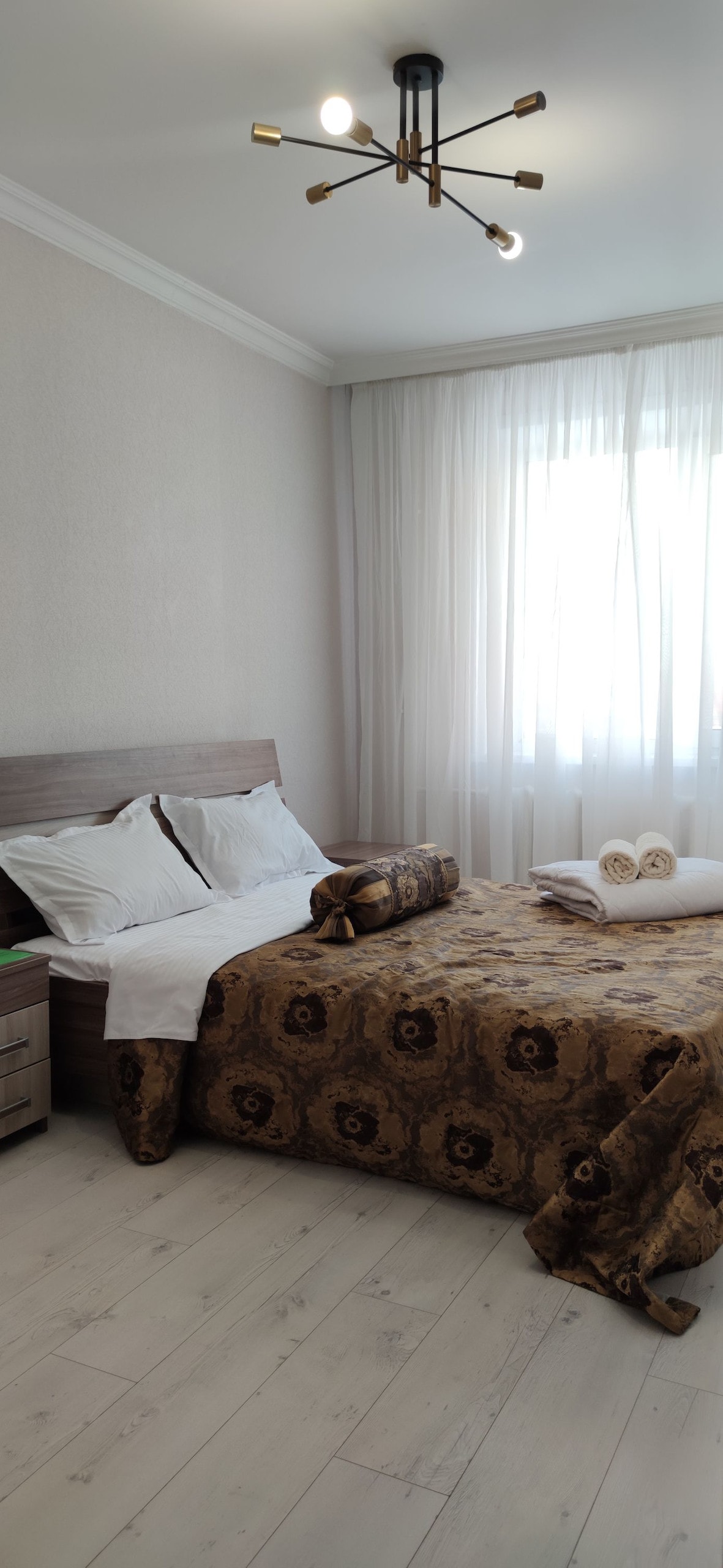 A bright bedroom features a spacious bed adorned with a decorative brown and gold bedspread. Large windows with sheer white curtains allow natural light to fill the room. Soft towels are neatly arranged at the foot of the bed, complemented by a bedside table with storage.