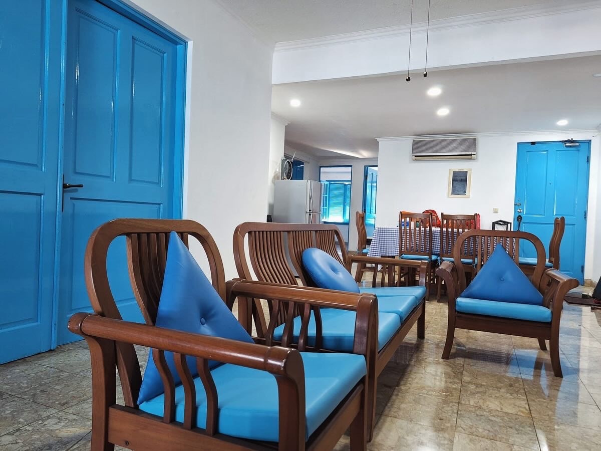 A welcoming common area is featured, showcasing wooden seating with blue cushions arranged for relaxation. The space is brightened by natural light, with blue doors and walls providing a vibrant contrast. A dining area and kitchen appliances are visible in the background.