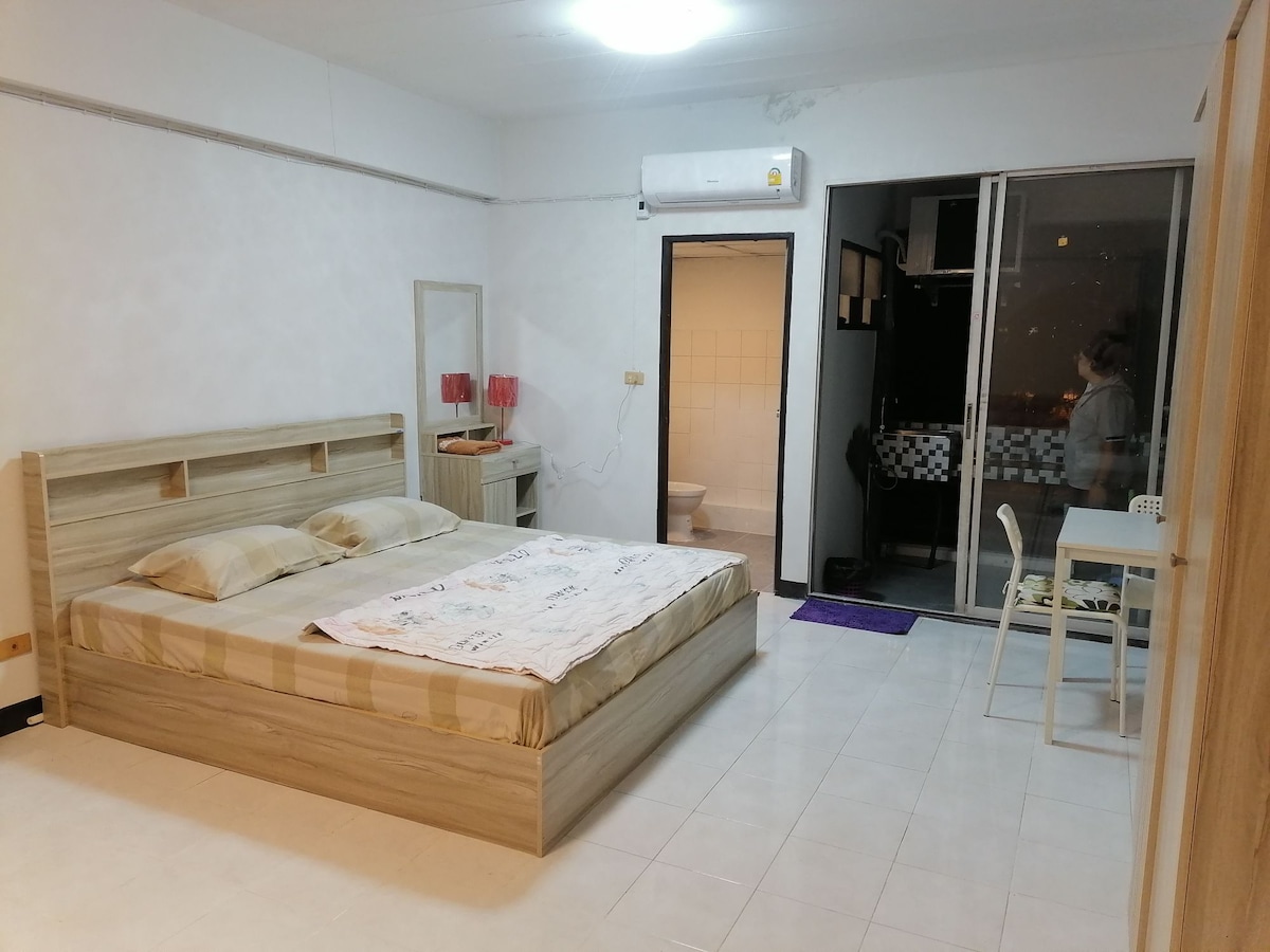 A spacious room is featured, containing a large wooden bed with light-colored bedding. A mirror and soft lighting accompany the bed, while a small dining table is positioned nearby. A door leads to a kitchenette area, and large glass doors provide access to the balcony.