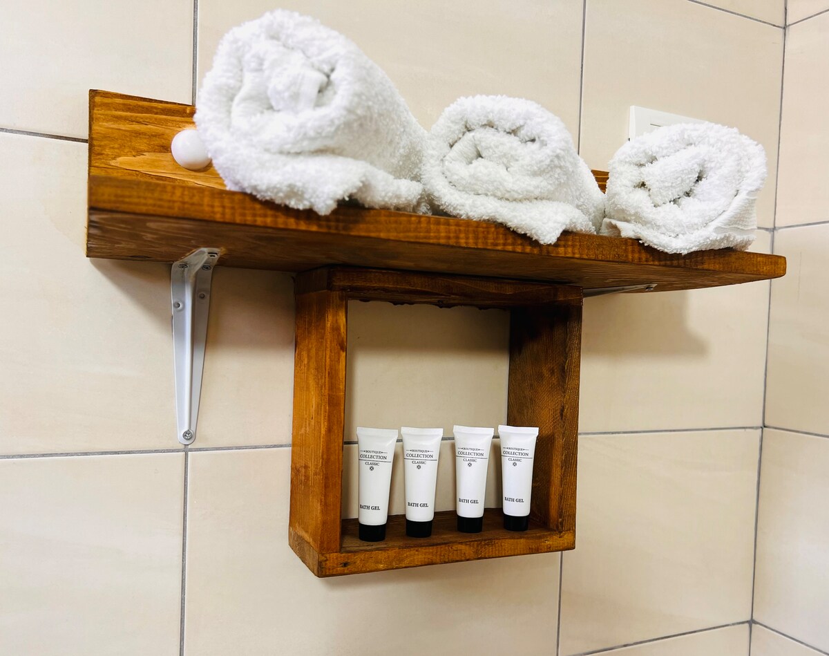 A wooden shelf is mounted on a bathroom wall, displaying three neatly rolled white towels and five small tubes of toiletries arranged below. The tiles in the background feature a neutral tone, creating a clean and organized appearance.