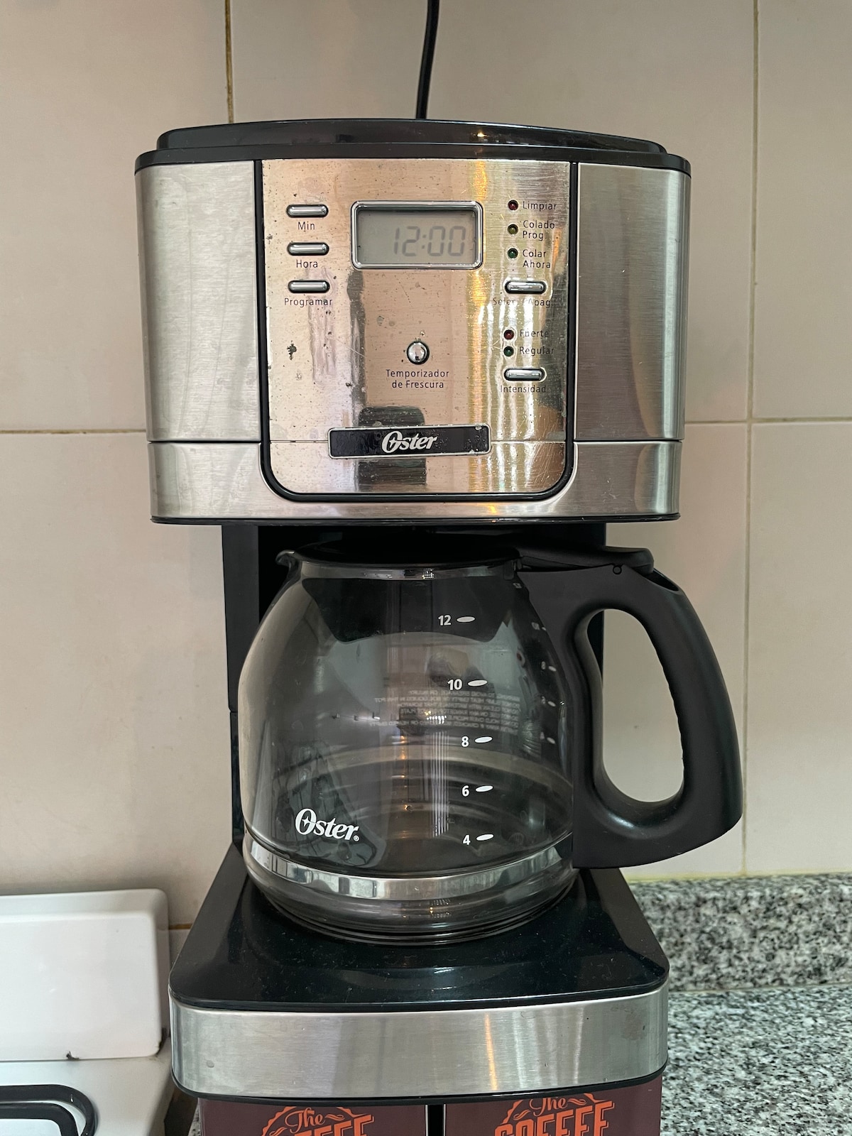 A coffee maker is displayed on a kitchen countertop, featuring a stainless steel body and a glass carafe. The digital control panel shows the time, and the carafe has measurement markings along the side, indicating its capacity.