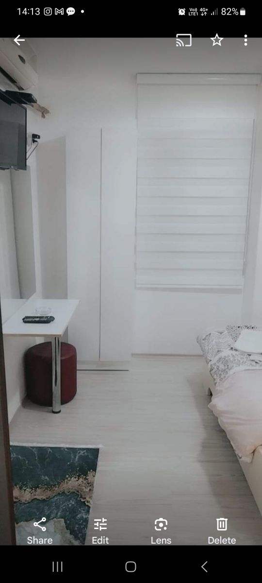 A compact bedroom is presented with a spacious bed covered in a light-colored blanket. A flat-screen television is mounted on the wall, and a small table with a chair is positioned beside a mirrored wardrobe. Natural light filters through a partially closed window.