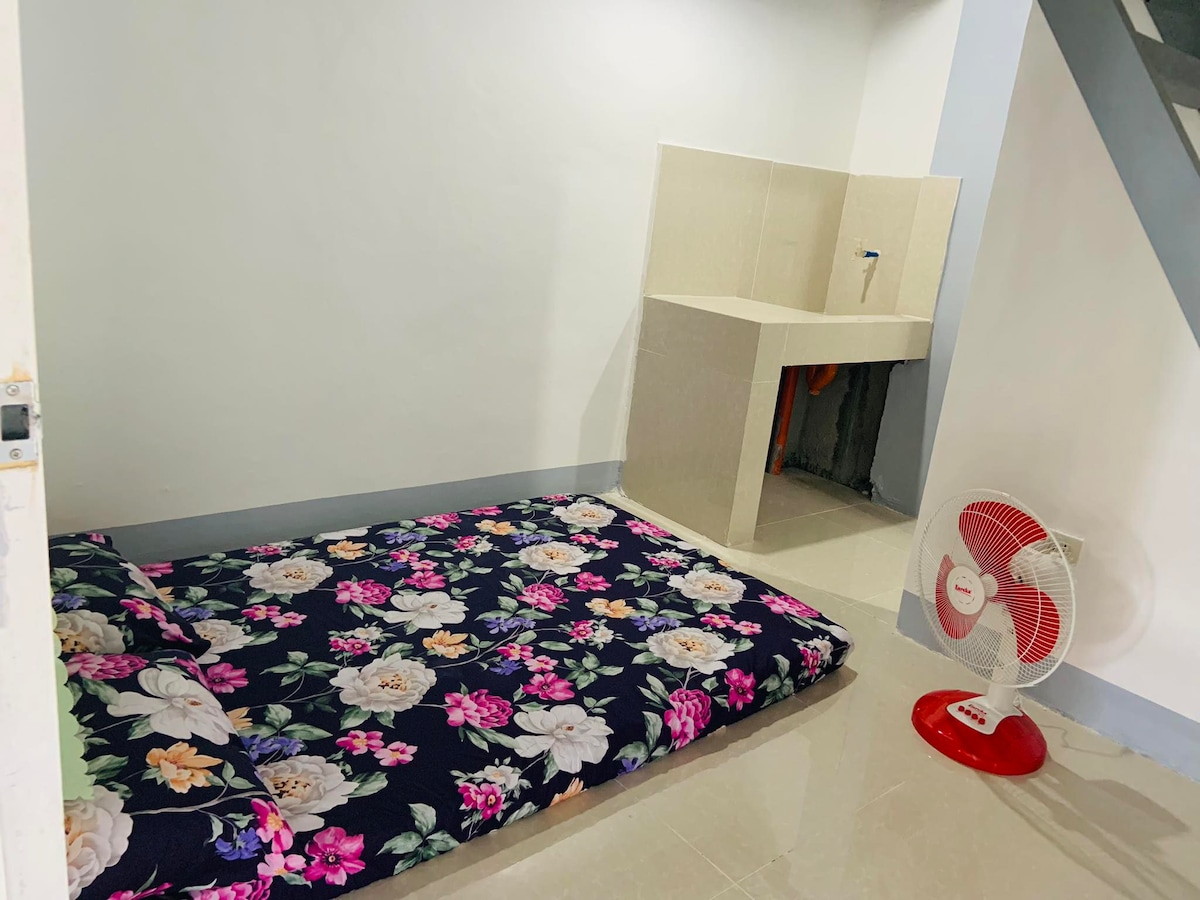A sleeping area features a floral-patterned mattress placed on the floor, complemented by a standing fan nearby. A simple countertop with a faucet is visible against the wall, contributing to a functional and minimalist design.