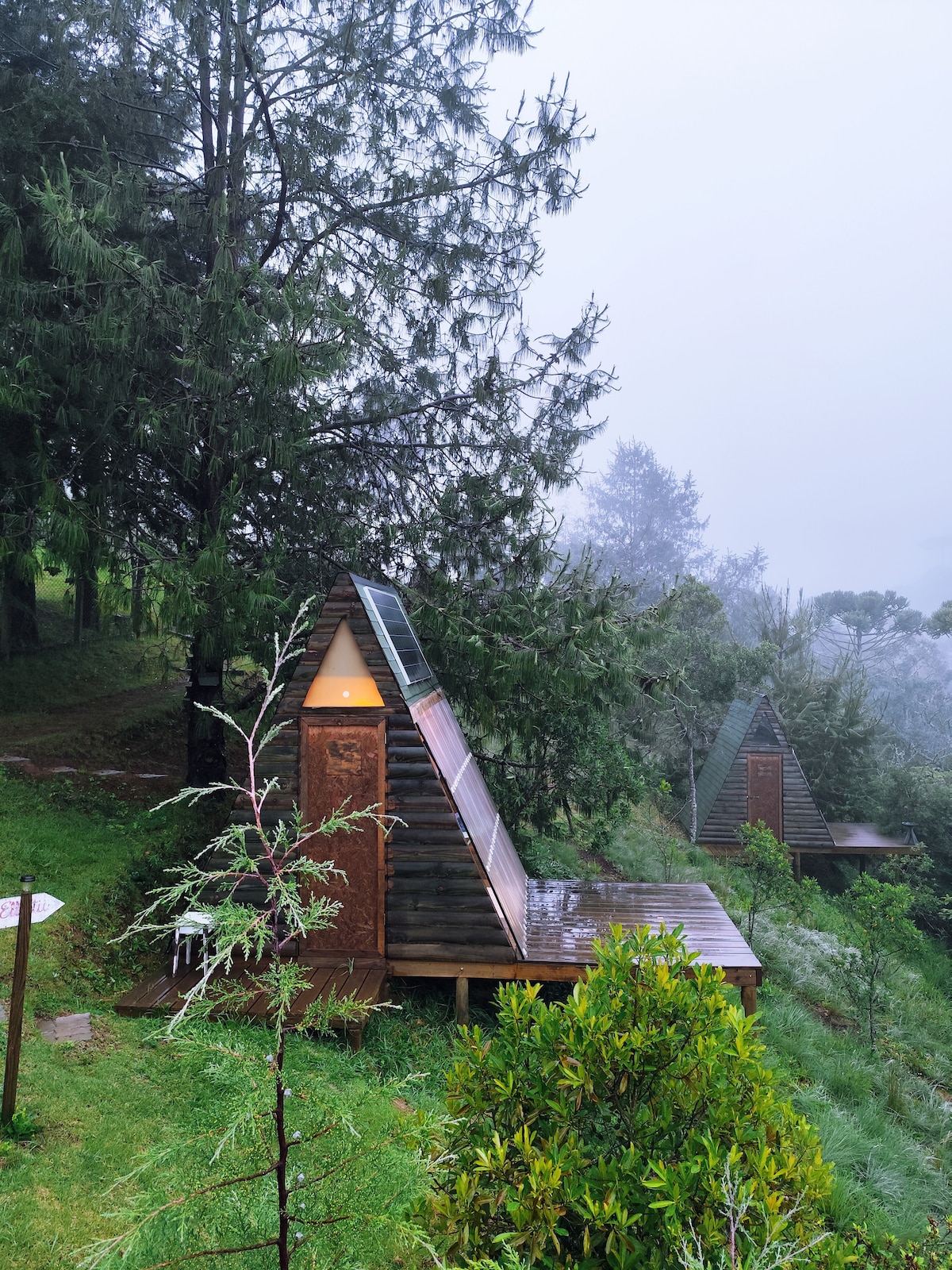 Two A-frame cabins are nestled among tall trees, surrounded by lush greenery. Each cabin features a triangular roof and wooden exterior, with a deck overlooking the serene landscape. A misty atmosphere enhances the tranquil setting.