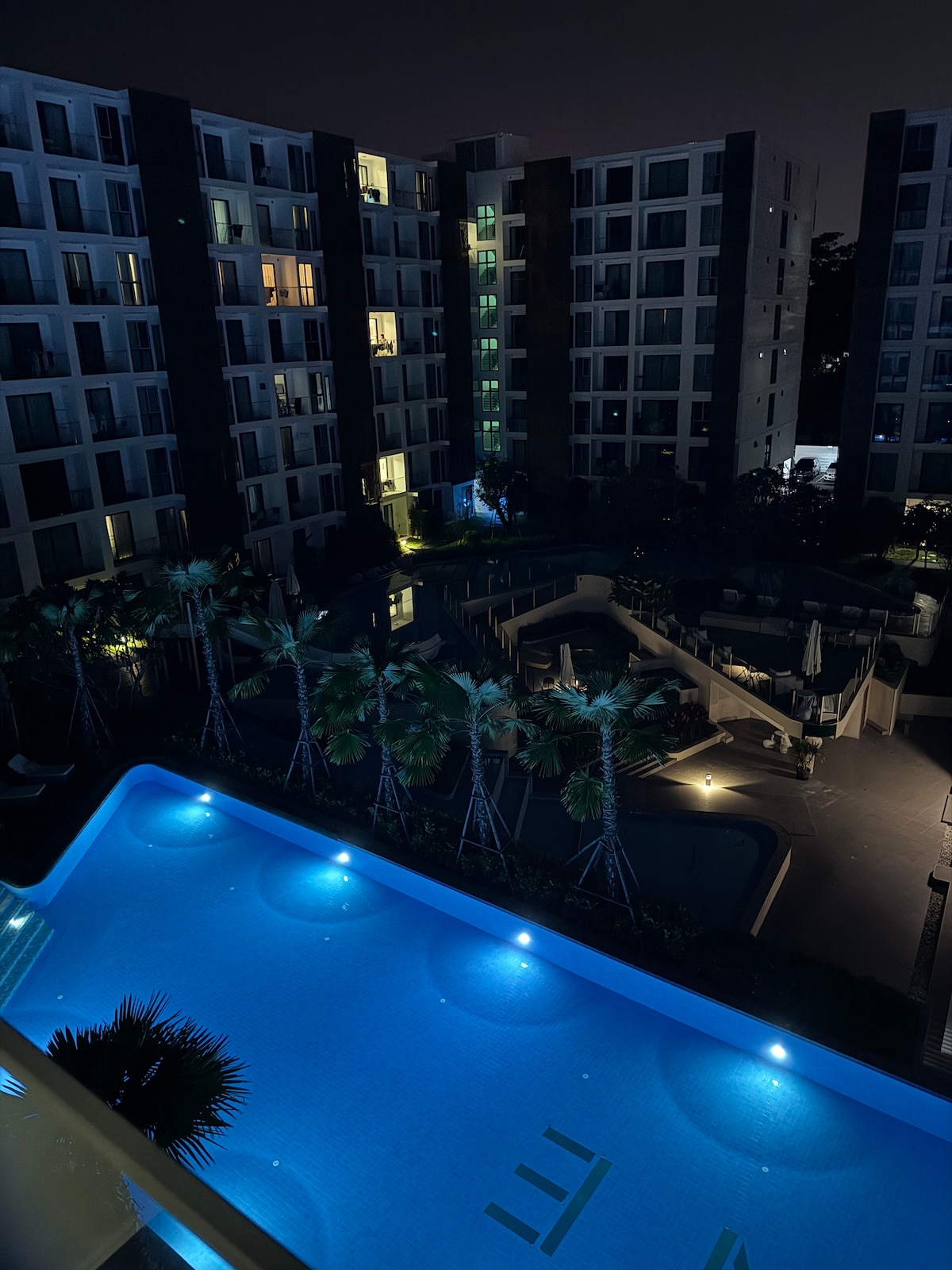 A night view showcases the expansive swimming pool illuminated in soft blue lights. Surrounding buildings feature illuminated windows, and tropical palm trees are lined along the pool area, enhancing the serene atmosphere of the space.