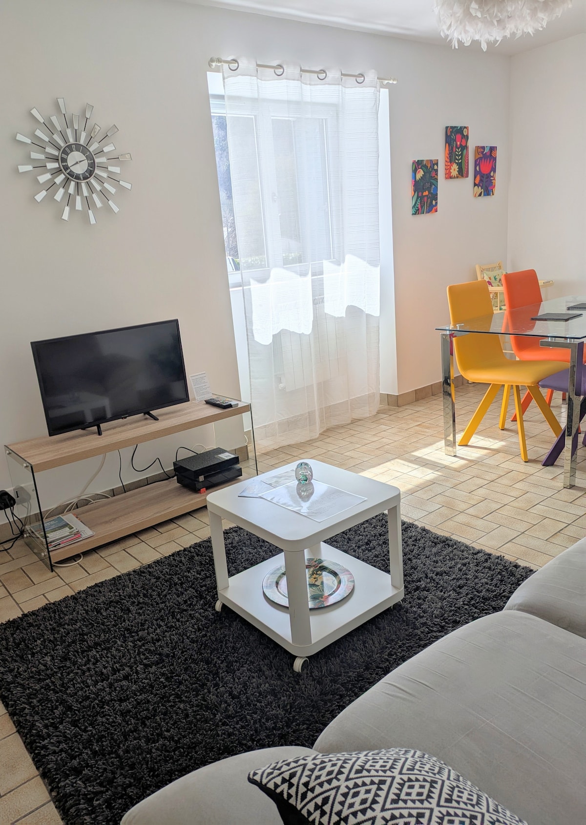 A living area is displayed, featuring a flat-screen TV and a light-colored sofa on a dark rug. A coffee table on wheels is positioned in front of the sofa. Colorful chairs surround a glass dining table, and natural light enters through sheer curtains.