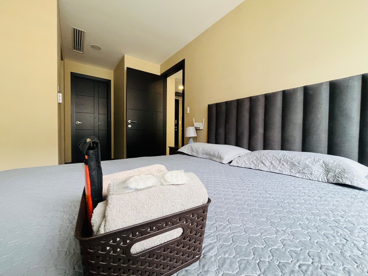 A spacious sleeping area features a queen-sized bed with a textured gray coverlet. Soft towels are neatly arranged in a basket on the bed. Dark doors are visible in the background, leading to the room entrance.