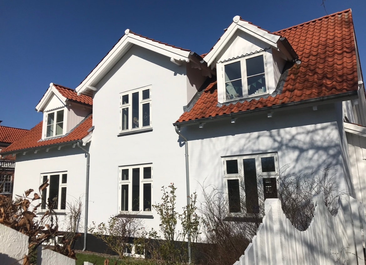 A two-story house features a combination of white exterior walls and a red-tiled roof. Multiple windows are present, allowing natural light to fill the interior. Surrounding greenery adds a touch of nature to the property, creating a cheerful and inviting exterior.