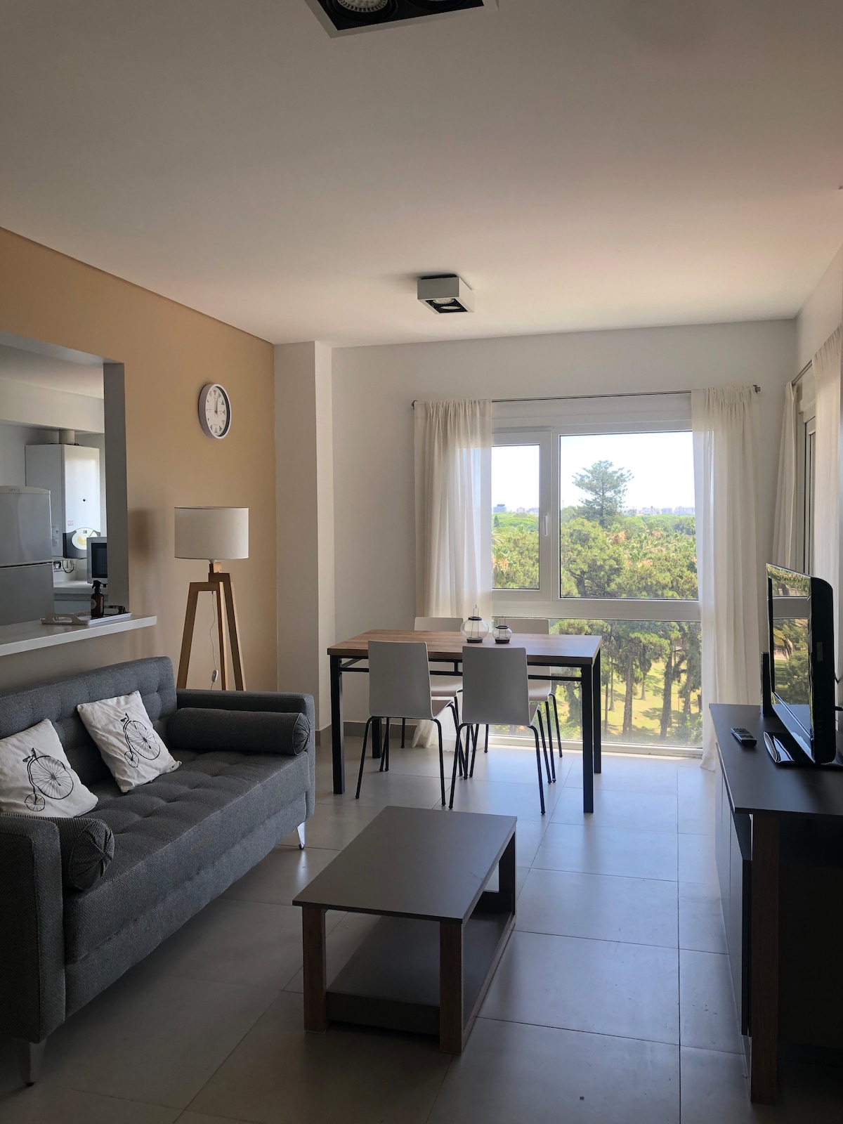 A bright living area features a grey sofa facing a flat-screen TV and a light wood coffee table. A dining table with chairs is positioned near a large window, allowing natural light to illuminate the space and providing a view of greenery outside.