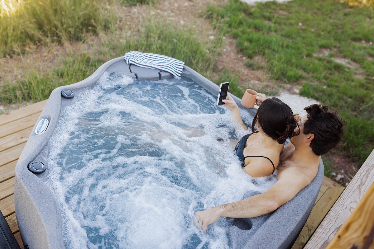 A hot tub is set on a wooden deck, surrounded by greenery. Two individuals relax together, one holding a smartphone and the other a mug. Soft bubbles rise from the water, creating a serene atmosphere in the natural setting.