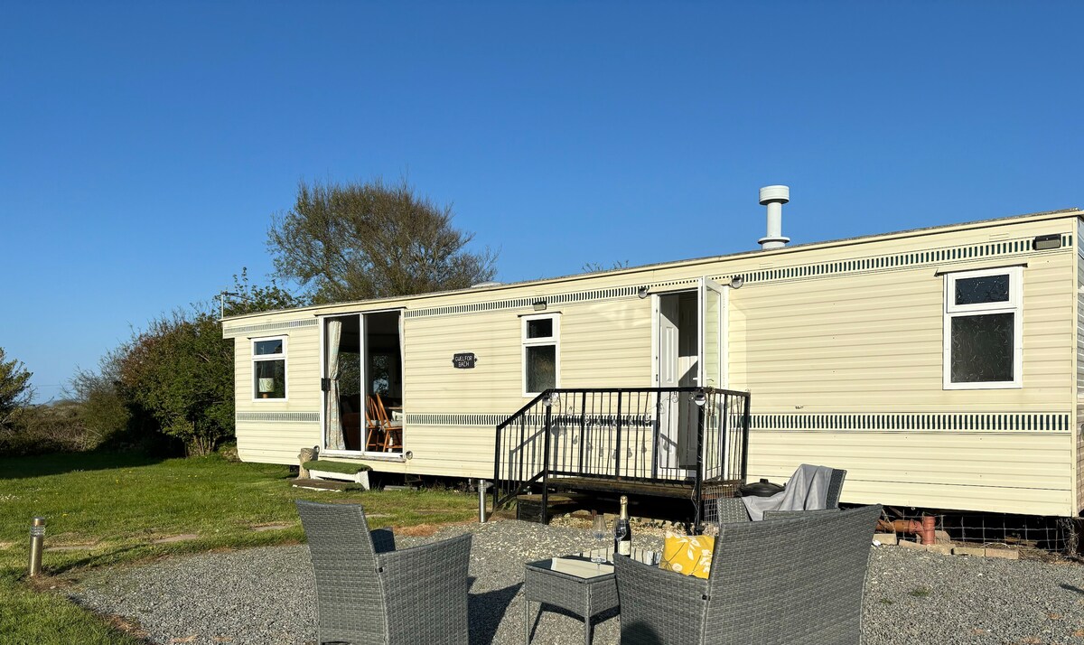 A static caravan is depicted with a welcoming entryway featuring steps leading up to the door. Two outdoor chairs are positioned nearby on a gravel area, surrounded by greenery under a clear blue sky.