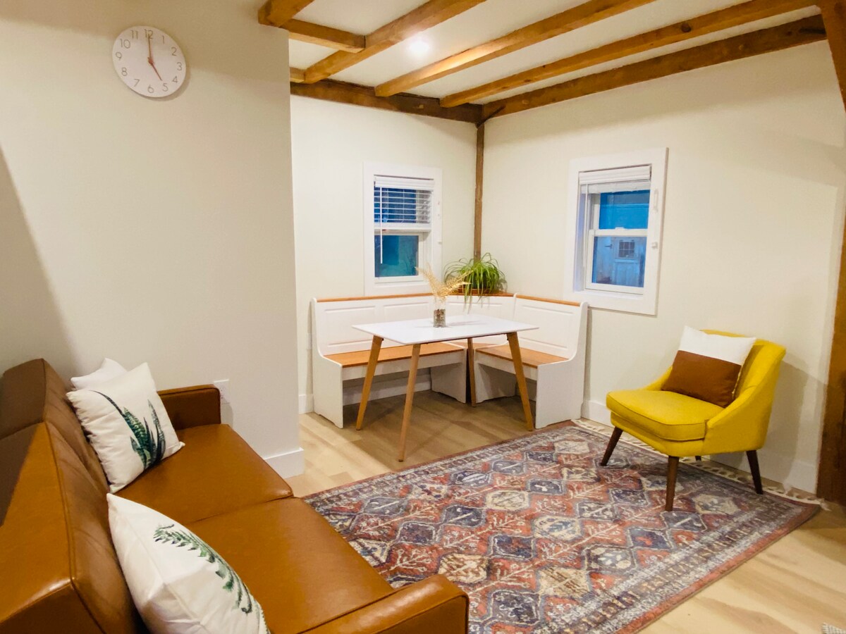 A cozy living space features a brown leather couch and a bright yellow accent chair. A wooden dining table with built-in bench seating is present, complemented by an area rug with intricate patterns. Natural light enters through the windows, highlighting the beams on the ceiling.
