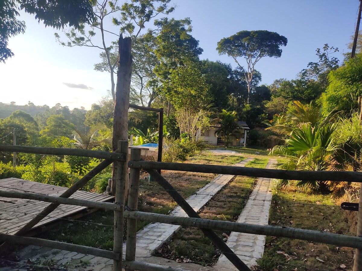 A rustic wooden gate opens to a gravel path leading through lush greenery. The scene is framed by tall trees and tropical plants that create a serene ambiance. In the distance, a cozy structure is partially visible, suggesting a peaceful retreat in nature.