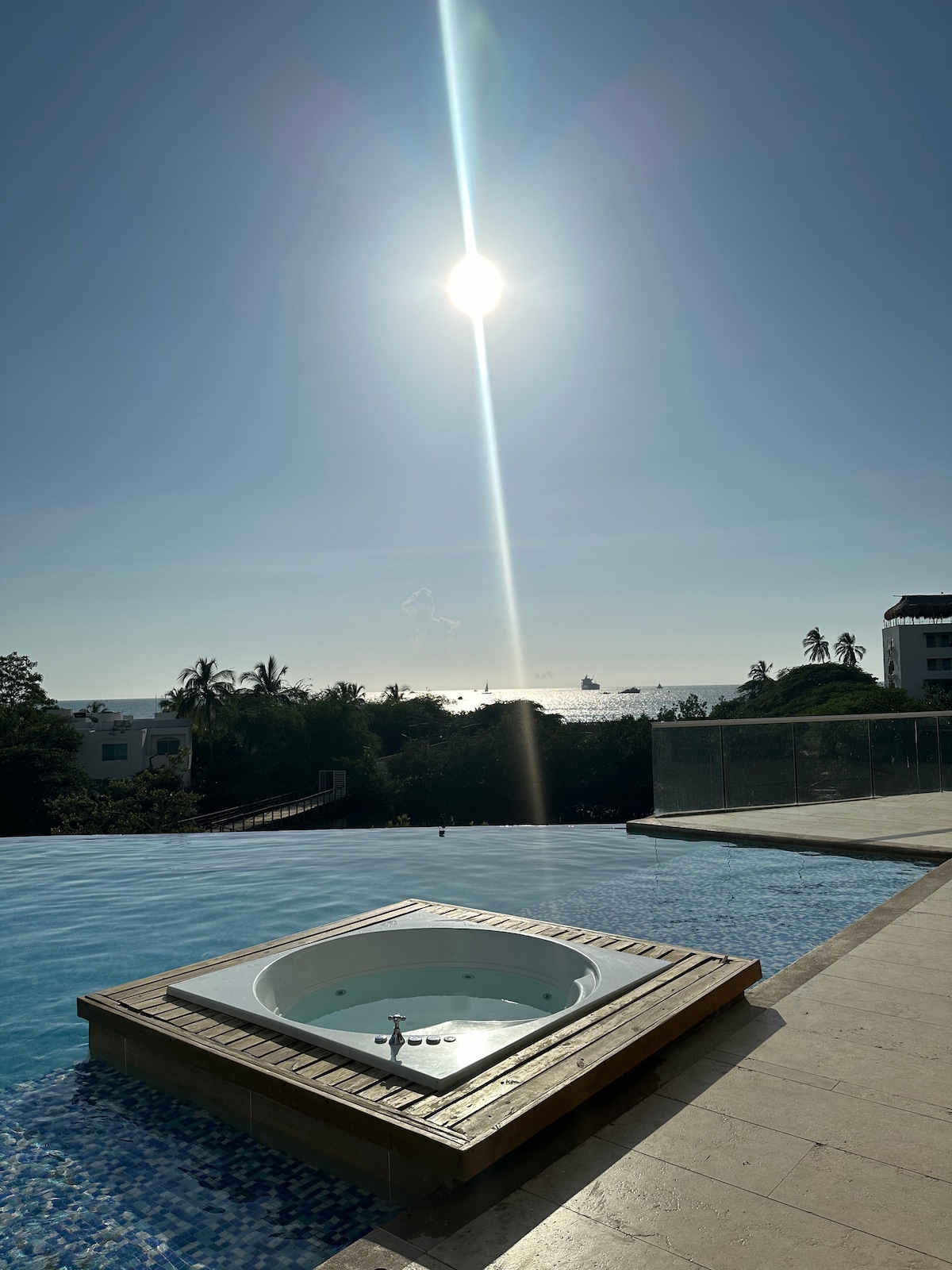 A serene scene showcases a jacuzzi placed on a wooden deck by the edge of an infinity pool. The sun reflects off the water, creating a bright contrast against the clear sky. Lush greenery and hills are visible in the background.