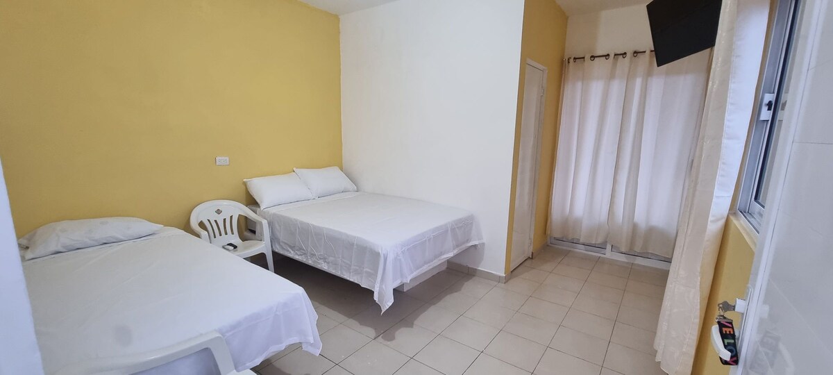 The room features two spacious beds with white linens, complemented by light yellow walls. A plastic chair is positioned near the wall, while curtains allow natural light to enter. A window is partially visible, and a television is mounted above.