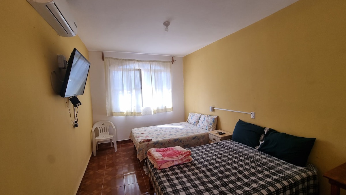 A cozy sleeping area is presented with two beds, one featuring floral bedding and the other adorned with a checkered pattern. A flat-screen TV is mounted on the wall, and a window is dressed with light curtains, allowing natural light to fill the space.
