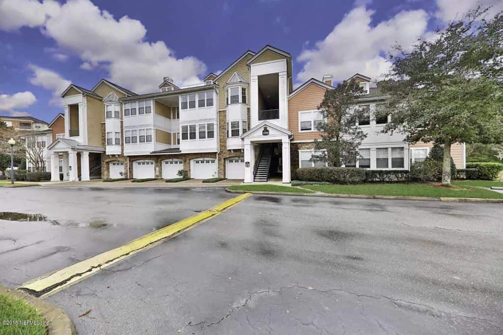 The exterior of a multi-story building is presented, featuring a blend of beige and white siding. The entrance is marked by a covered porch and decorative lighting. Lush greenery surrounds the structure, with a paved pathway leading to the front. Garage doors are visible on the ground level.