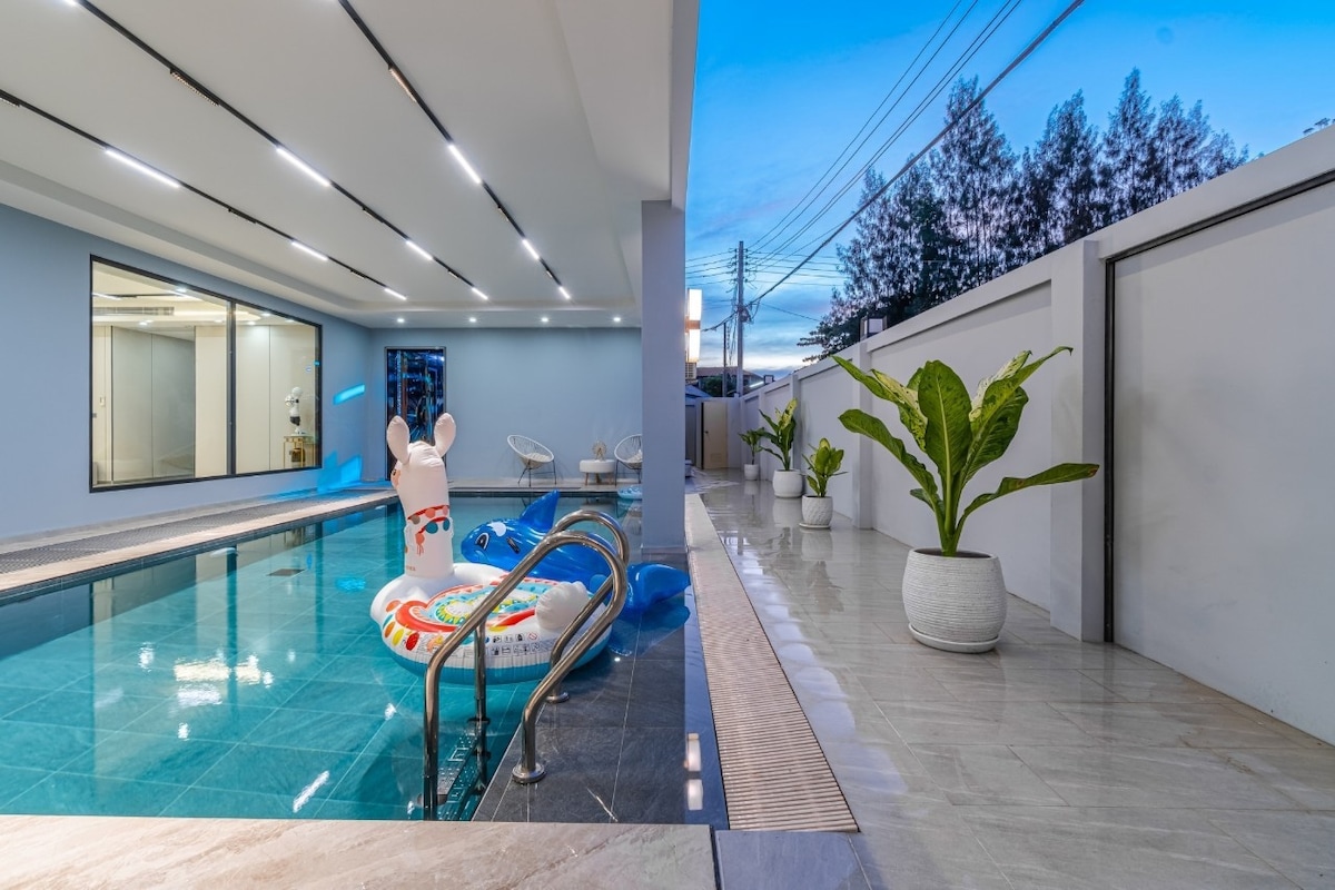 An outdoor swimming pool is surrounded by sleek tiles, featuring a colorful inflatable float sitting on the water. Large windows provide a view into the adjacent indoor area, while potted plants add greenery to the space. The area is well-lit under a soft blue sky.