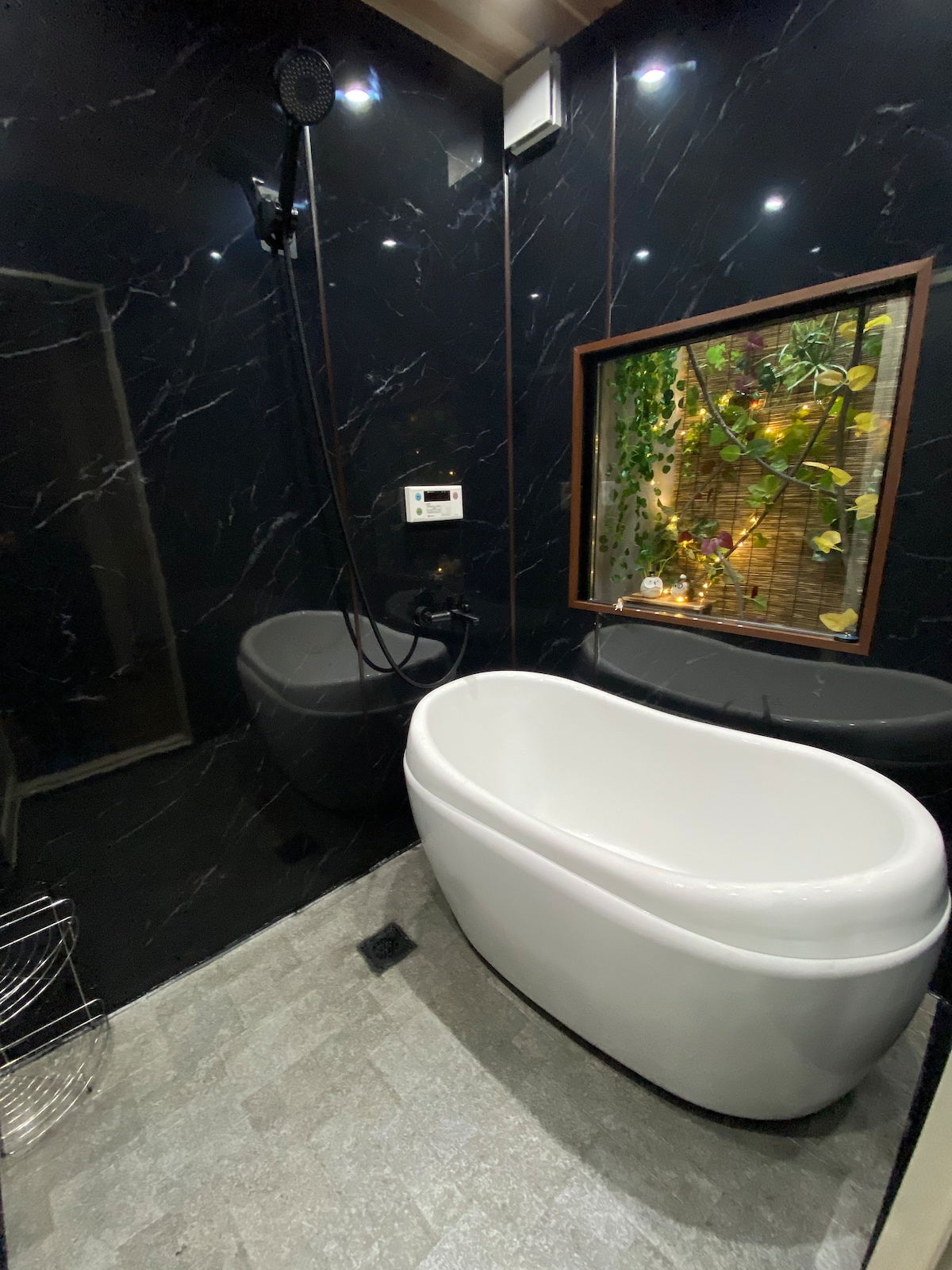 A modern bathroom features an elegant freestanding bathtub and a sleek shower area. Dark marble walls create a striking contrast with the white tub. A window showcases a glimpse of greenery and warm lighting, enhancing the tranquil ambiance of the space.
