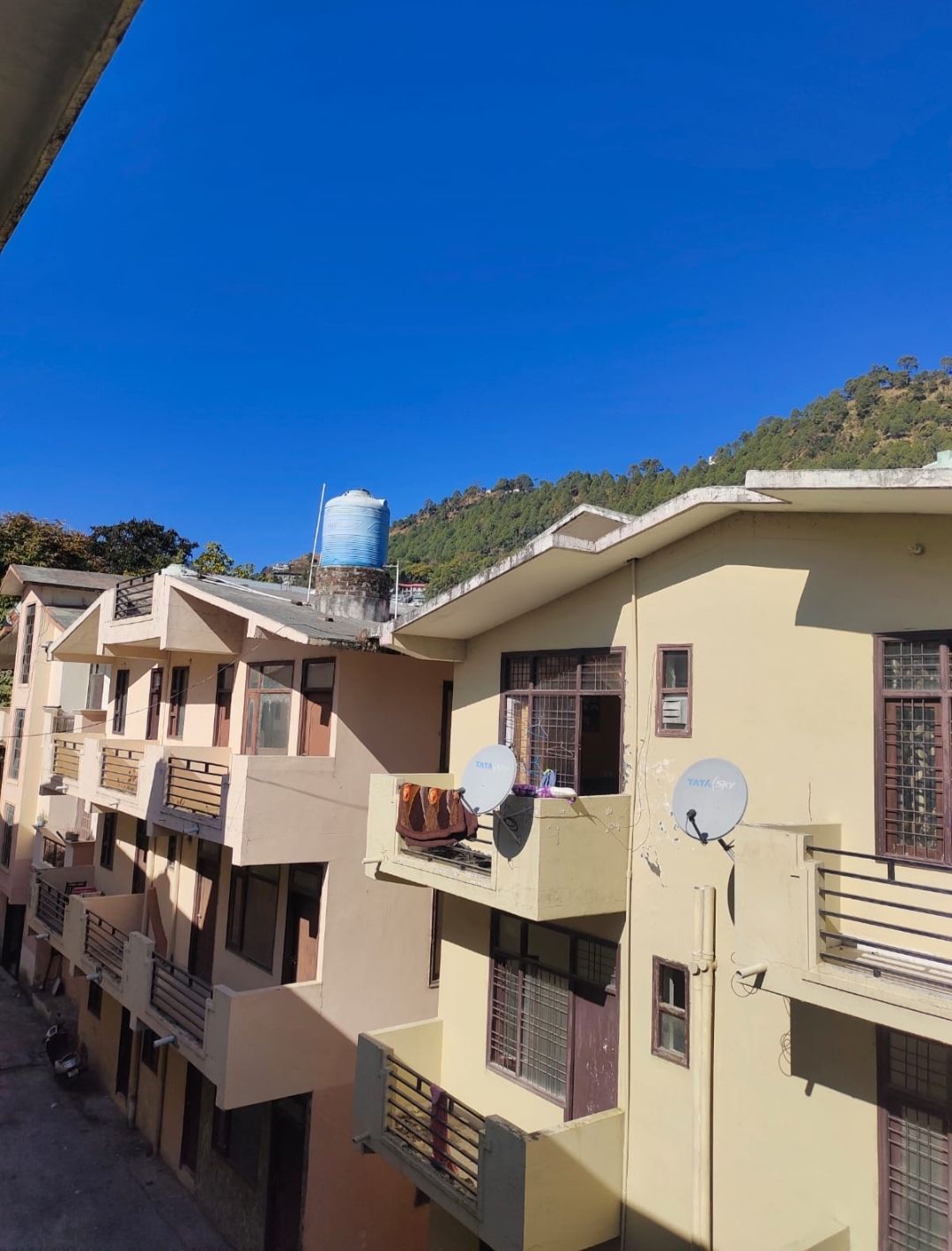 The image depicts a view of a multi-story building facade under a clear blue sky. Satellite dishes are mounted on some balconies, and a water tank is visible on the rooftop. Lush greenery is seen on the hillside in the background.