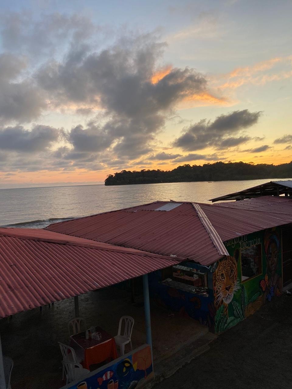 A vibrant sunset paints the sky above the coastline, reflecting on the calm waters. A rustic building with a red corrugated roof and colorful murals is partially visible in the foreground. Lush greenery can be seen on the distant shore, enhancing the natural beauty.