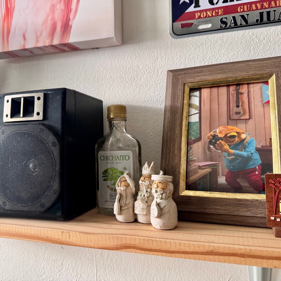 A wooden shelf is adorned with a speaker, a bottle of Chichaito, and a framed photo depicting a figure with a frog mask. Three small ceramic figures are positioned together, showcasing unique, hand-crafted details.