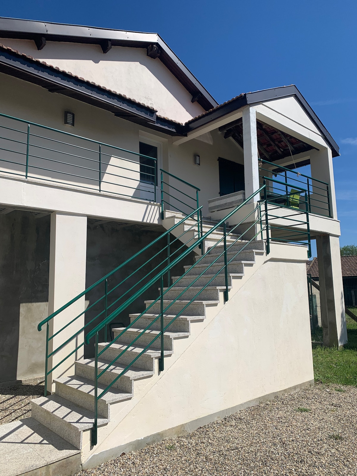 A set of concrete stairs leads to an entrance, framed by a light-colored exterior wall. Green railings provide support along both sides. The upper level is partially shaded, with a glimpse of a sunny sky in the background, creating a pleasant approach to the home.