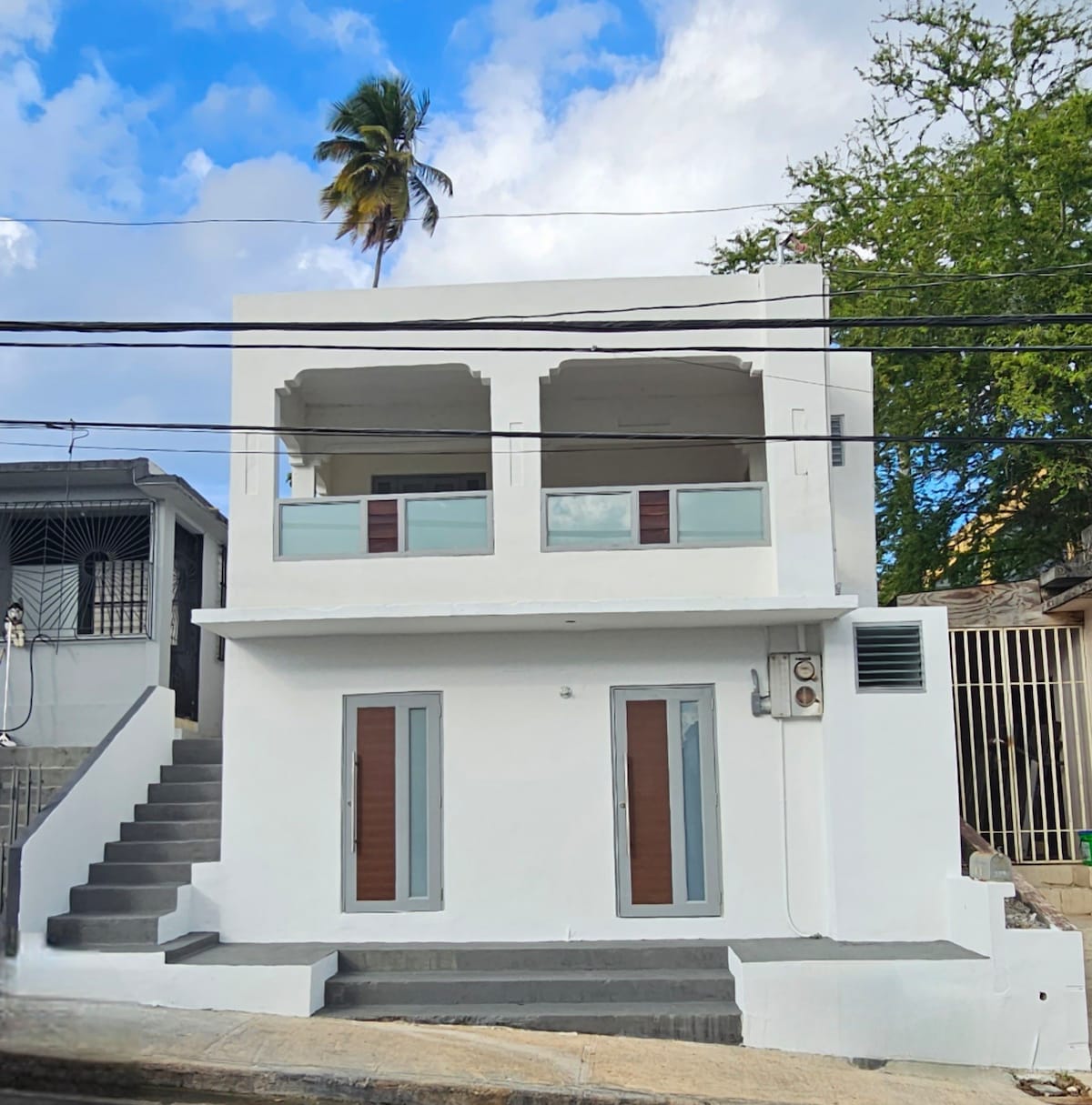A white two-story building is depicted, featuring a set of steps leading to a first-floor entrance. Two large balconies with glass railings are visible above, complemented by two doorways on the ground level. The surroundings include palm trees and nearby structures.