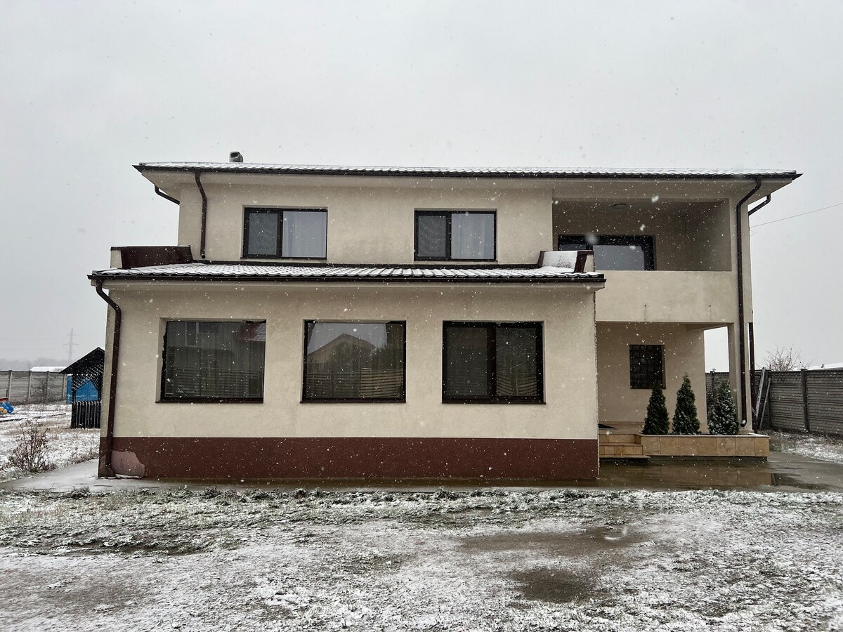 The exterior of a modern two-story villa is shown, featuring a light-colored facade with large windows. Snow gently falls on the property, creating a serene winter scene. The entrance is complemented by steps leading to a covered porch, surrounded by small evergreen plants.