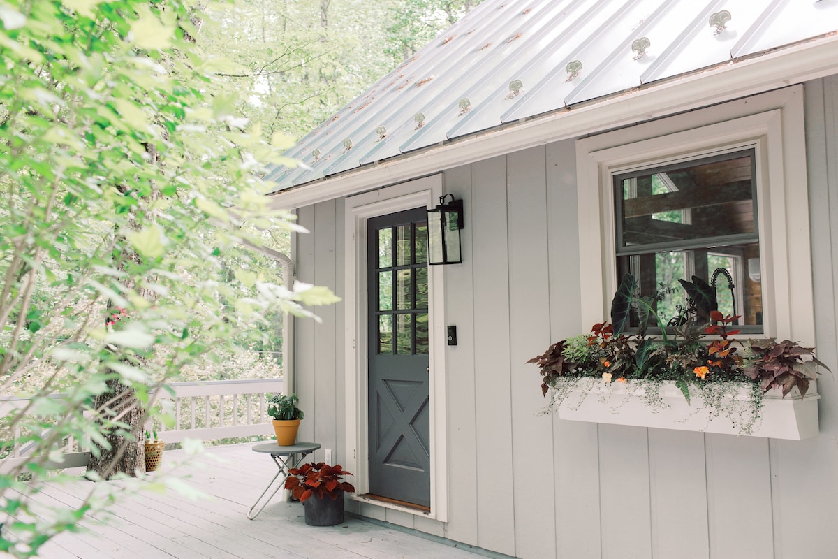 The exterior entrance showcases a charming door flanked by a planter box filled with vibrant flowers. A small table stands on the porch, complemented by lush greenery surrounding the cabin. Natural light enhances the serenity of the welcoming space beneath the metal roof.