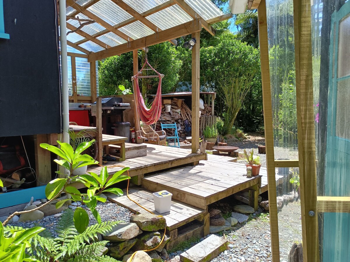 A wooden deck is visible, connected to a covered area with a colorful hammock. Potted plants are seen alongside the deck, while a stack of firewood and additional seating can be observed in the background, surrounded by greenery.