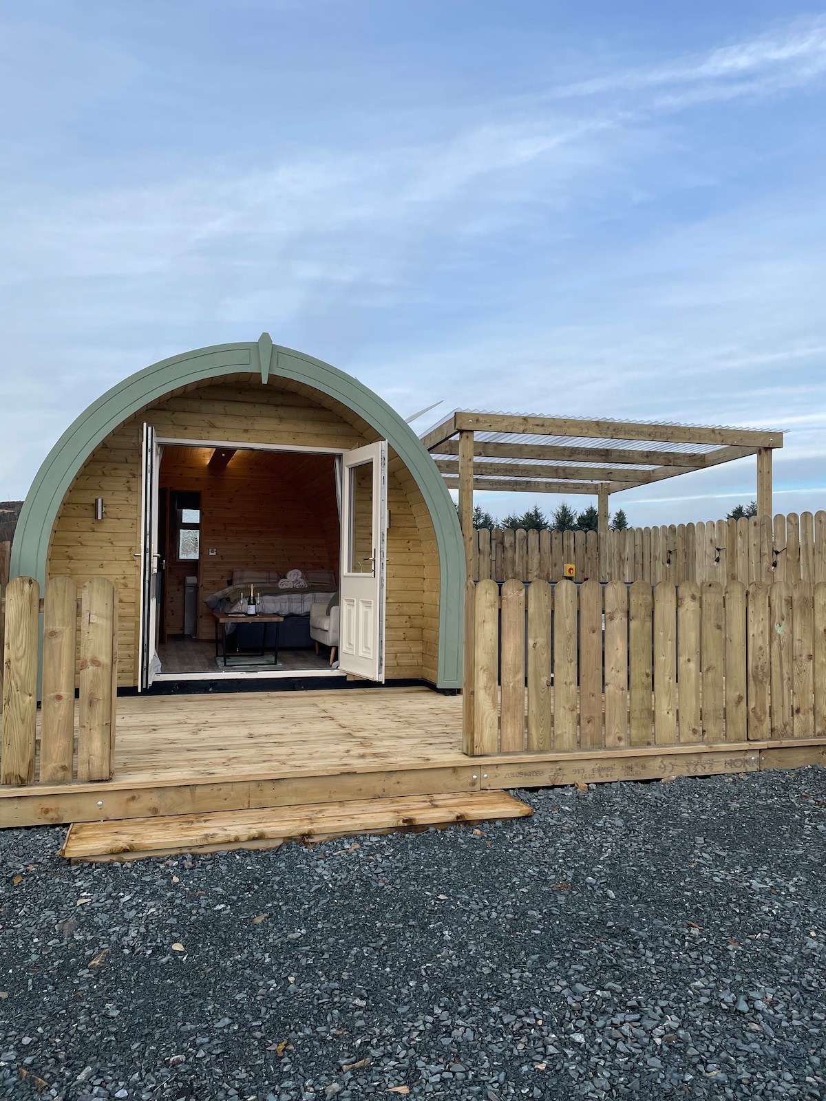 The exterior of a luxury pod is shown, featuring an inviting arched entrance. A wooden deck leads to a fenced area, with a simple roof structure above the entrance providing shelter. The pod's natural wood exterior blends seamlessly into the surroundings.