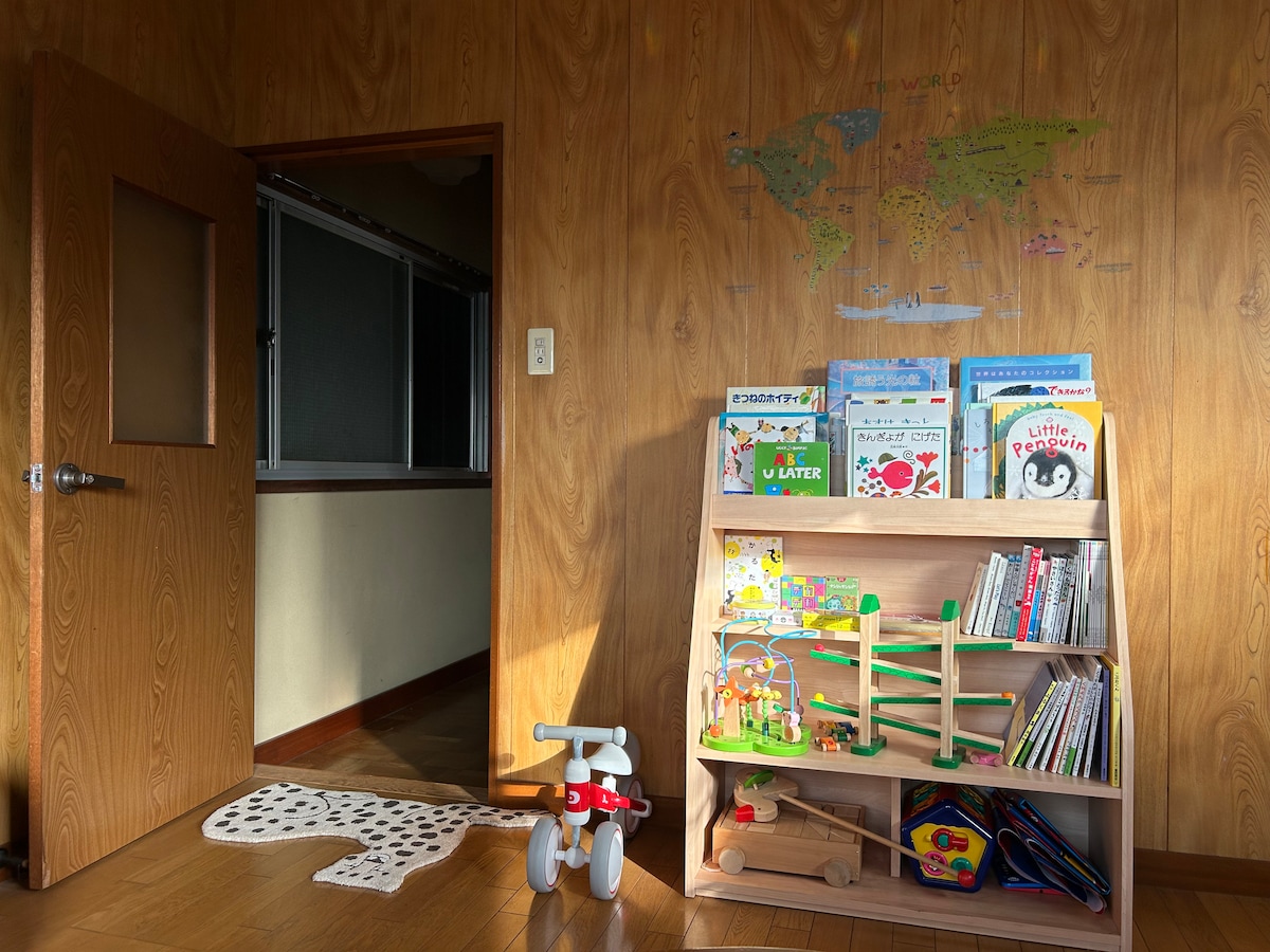 A brightly lit room showcases a wooden bookcase filled with children's books and toys. A toy tricycle and a soft area rug provide playful elements. The wall features a world map, and a door leads to another area of the home.